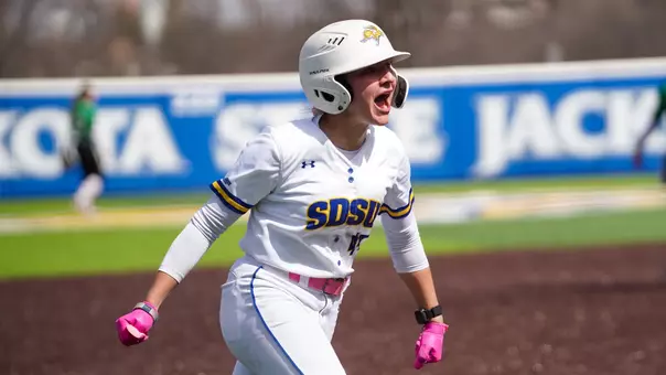 A woman in a white softball uniform runs towards home plate and yells enthusiastically