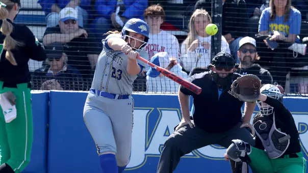 A woman in a grey uniform swings a bat and makes contact with a softball while a woman in black and green looks on