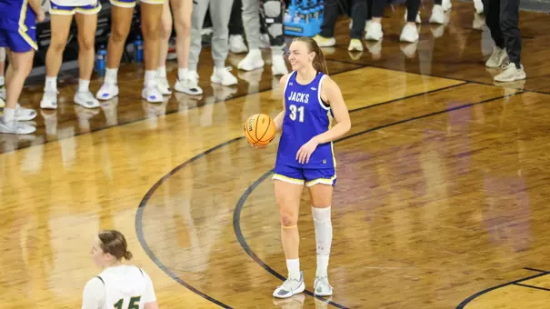 A women's basketball player in a blue uniform holds the ball and smiles while standing on the court during a game.