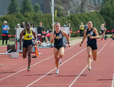 A female track athlete in black sprints down the front of the track in the 100-meter race with a compeitor to her right and a teammate to her left