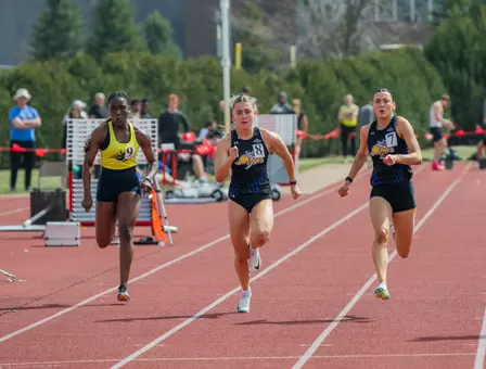 A female track athlete in black sprints down the front of the track in the 100-meter race with a compeitor to her right and a teammate to her left