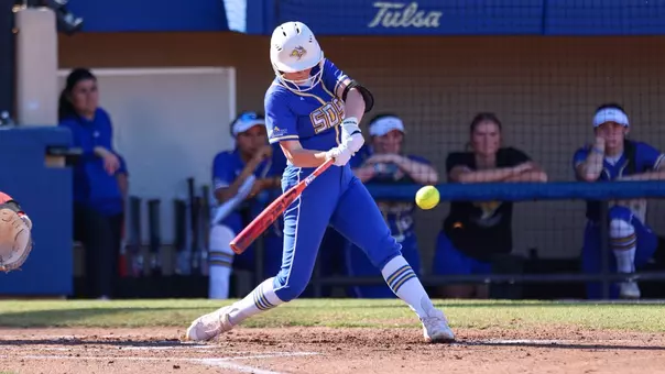 A woman in a blue softball uniform swings a bat and makes contact with a softball
