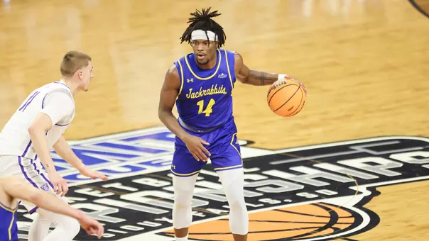 A man in a blue uniform dribbles a basketball with a defender in front of him