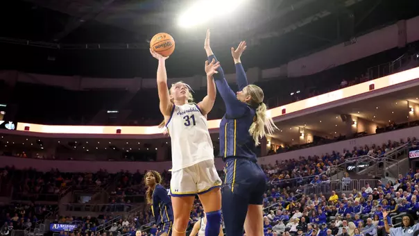 A women's basketball player in a white uniform extends her arms overhead and attempts a shot over a defender in a blue uniform.