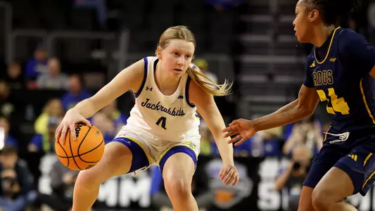 A women's basketball player in a white uniform dribbles on the basketball court as an opposing player in a navy uniform defends