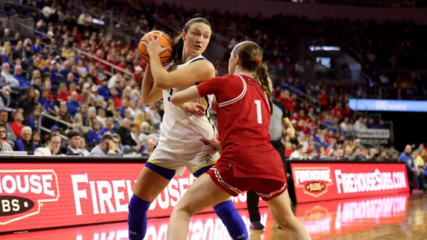 A player in a white uniform holds the basketball near the sideline while a defender in a red uniform extends an arm to contest the play on the court, with a packed arena crowd in the background.