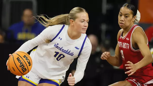 A women's basketball player in a white uniform handles the basketball as a player in a red uniform defends