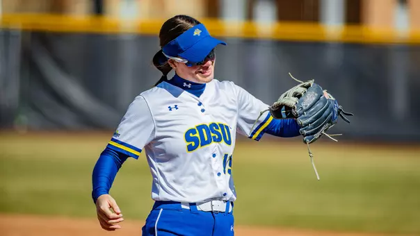 A softball player wearing an SDSU uniform stands on the field holding a glove out to the side, with a dirt infield and outfield fence visible in the background.