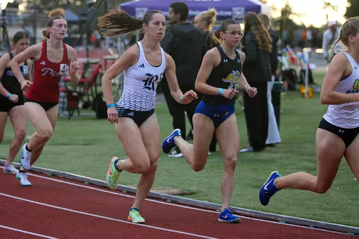 A female runner races around the curve of the track in a black and blue uniform as a white uniformed competitior tries to pass her