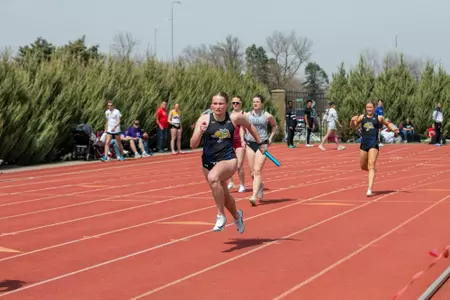 A female track athlete in a black uniform races down the backside of the track after taking the baton from her teammate, who is seen in the background slowing her strides.