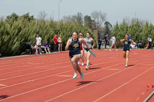 A female track athlete in a black uniform races down the backside of the track after taking the baton from her teammate, who is seen in the background slowing her strides.