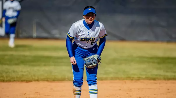 A woman in a white jersey and blue pants readies for a pitch while playing third base on a softball diamond