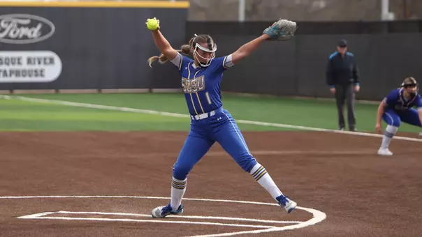 A woman in a blue softball uniform winds up to throw a pitch on a softball diamond