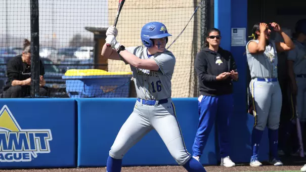 A woman in a grey softball uniform holds a bat as she prepares for a pitch