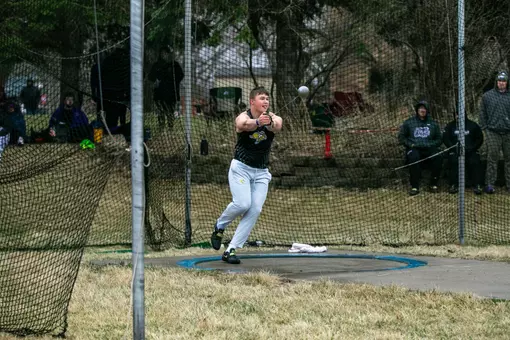 A male track and field thrower wearing a black jersey top and grey sweatpants begins spinning around in his hammer throw attempt
