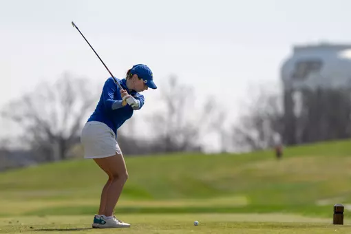 A women's golfer in a blue hat and white skirt swings through on the tee box with a golf club.