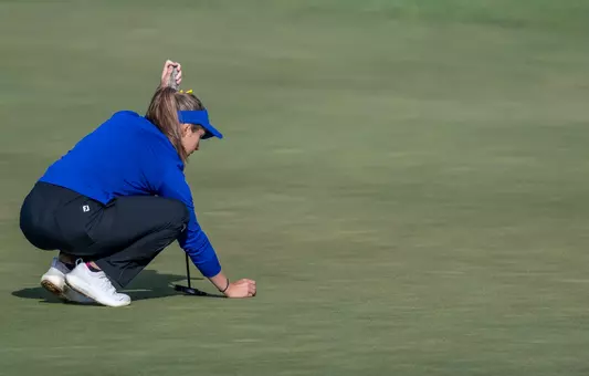 A women's golfer in a blue hat and blue long sleeve lines up her ball before putting.