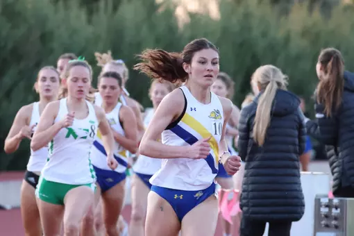 A female runner in a whilte jersey and blue shorts is depicted from the thighs up, rounding the track ahead of a blury pack of competition