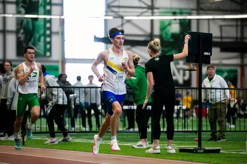 A male track athlete in a white jersey, blue shorts, and a blue headband, races around the curve of the track trailed by a competitor five steps behind him.