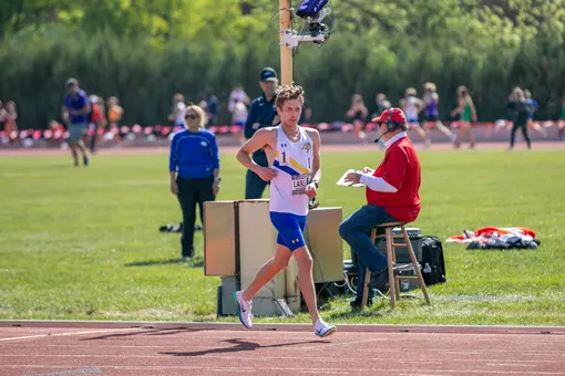 A male track athlete in a white jersey and blue shorts slows his stride after crossing the finish line