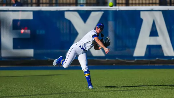 A woman in a white softball uniform throws a ball in the air and leans on her left leg during her follow through