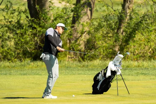 A men's golfer in a white hat swings his golf club with his bag nearby on the course.