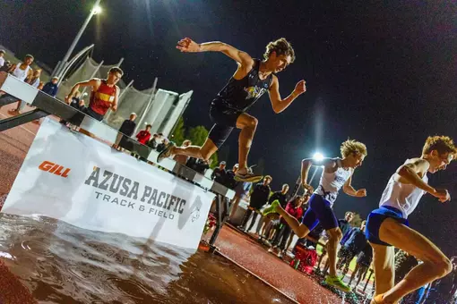 A male athlete is depicted in a black jersey and shorts, leaping over the steeplechase water barrier. The photo is rotated by roughly 45 degrees, putting the athlete at an angle.