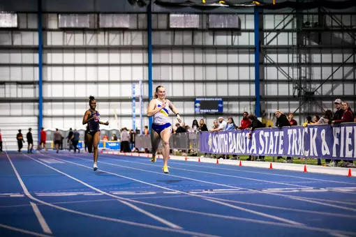 A female track athlete in white and blue sprints down the straight towards the finish line with a competitor in black several meters behind her