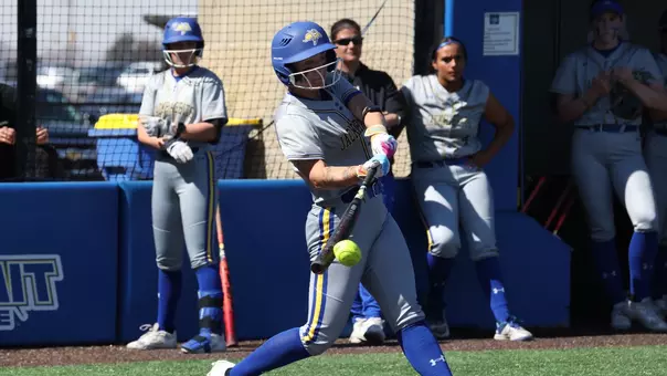 A woman in a grey softball uniform swings a bat and makes contact with a softball. Players and a coach on the same team look on from the dugout behind her.