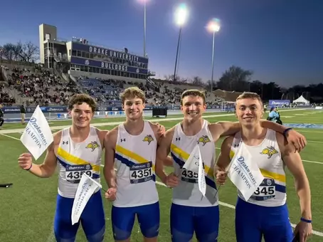 Four track athletes wearing white and blue uniforms pose of a photo with their arms around each other, each holding white Drake Relays Champions flags