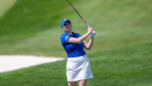 A women's golfer in a blue polo follows through on a swing and watches her ball on a golf course.