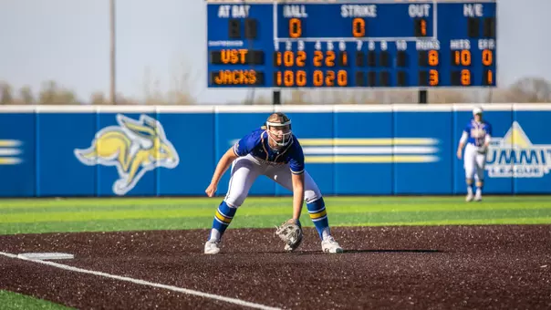 A softball player in a blue jersey playing third base crouches in a ready position, with her glove outstretched awaiting a hit from an opposing player. A turf field and scoreboard are visible.