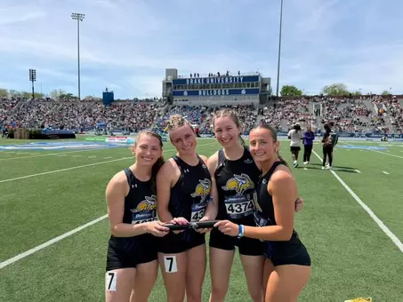 Four women in black track jerseys pose for a photo with each woman holding part of a black baton.