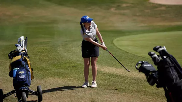 A women's golfer follows through on a shot on a golf course during competition. Two golf bags are visible at the edges of the frame.