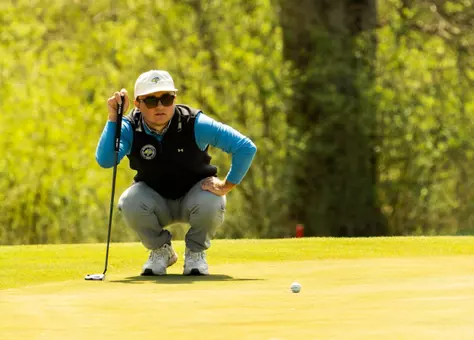 A men's golfer in a white hat and black vests kneels down to read the green and holding his club before attempting a put.