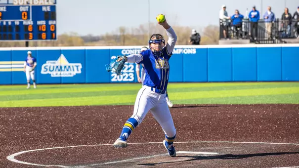 A woman in a blue jersey and white pants prepares to throw a softball from the circle on a softball diamond