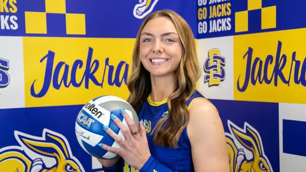 A South Dakota State volleyball student-athlete standing against a blue-and-yellow branded backdrop featuring the Jackrabbits logo, “Go Jacks,” and “Jackrabbits” text. The athlete wears a blue SDSU volleyball jersey and arm sleeve, holds a volleyball at chest level and faces the camera.