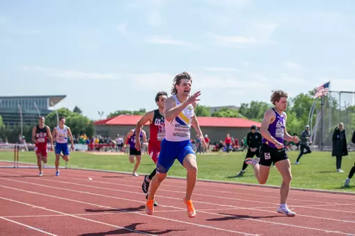 A male track athlete wearing a white and blue jersey races towards the finish line ahead of his competitors in purple and red.