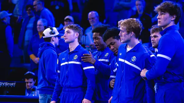 A wrestler in a blue tracksuit looks up during lineup intros before a dual.