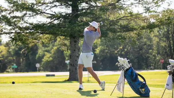 A men's golfer follows through on a swing during competition. The background features greenery on a golf course.
