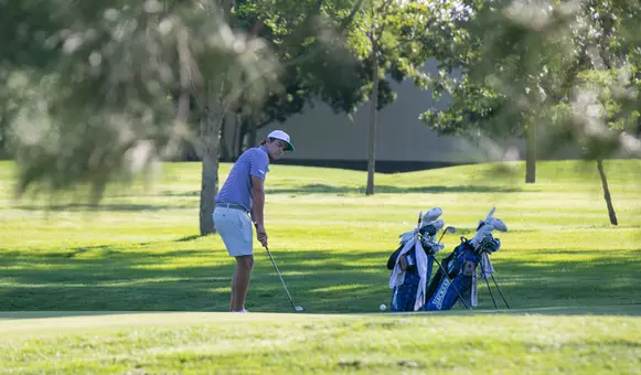 A men's golfer in a white hat and striped polo chips the ball on the green.