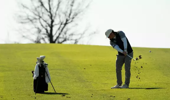 A men's golfer in a white hat and black vest swings through with his golf club kicking up the course.