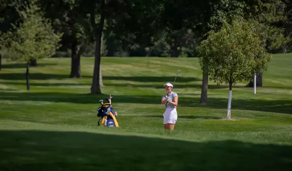 A women's golfer in a white hat and gray polo follows through on her chip shot to the green.