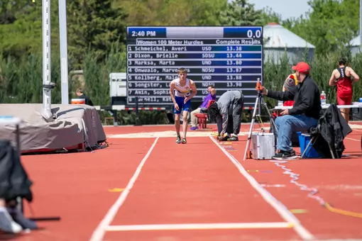 A male field athlete in a white and blue uniform gets into starting position at the end of the long jump runway with his torso bent and one leg behind him.