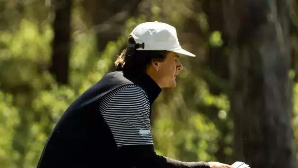 Golfer crouches low on a putting green, holding a putter and reading the line of a putt. The golfer wears a white cap, a dark vest over a striped long-sleeve shirt, light-colored pants, and golf shoes. The background shows trees and natural greenery in soft focus under daylight.