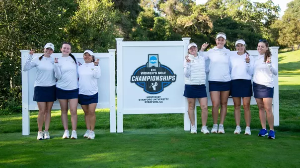 The SDSU women's golf team poses for a team photo in front of an NCAA golf championships sign. Seven women dressed in white tops, navy skirts and white hats stand in a line, each with one hand raised in a "bunny ears" signal.