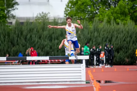 A male track athlete in a white jersey and blue shorts leaps off a steeplechase barrier into the water