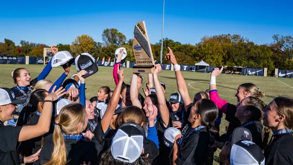 A group of women in black shirts jump up and cheer with a trophy lifted in the air by one woman