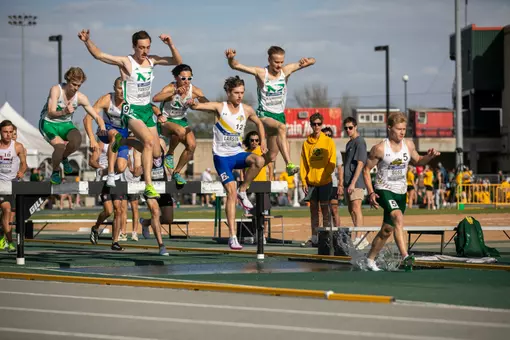 A male track athlete in white and blue is mid-fall into the water barrier of the steeplechase event