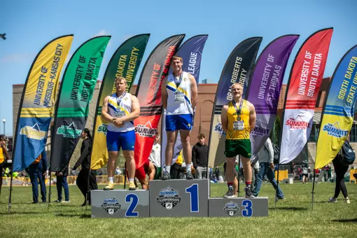 Three male throwers stand on a podium for the discus competition. The third-place finisher wears green and yellow, while first and second are teammates wearing white and blue.